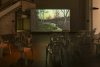 A large dark space shining on different mono bloc chairs positioned in front of a screen projection of a woodland at dawn or dusk.